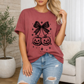A woman wearing a dusty rose short-sleeve t-shirt with a black graphic of two jack-o'-lanterns tied by a large bow, paired with distressed blue jeans in a cozy living room setting.