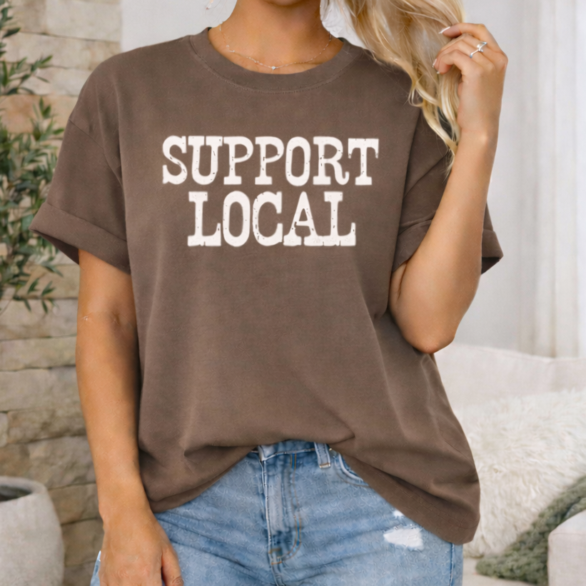 Person wearing a brown t-shirt with 'SUPPORT LOCAL' text in a casual setting.