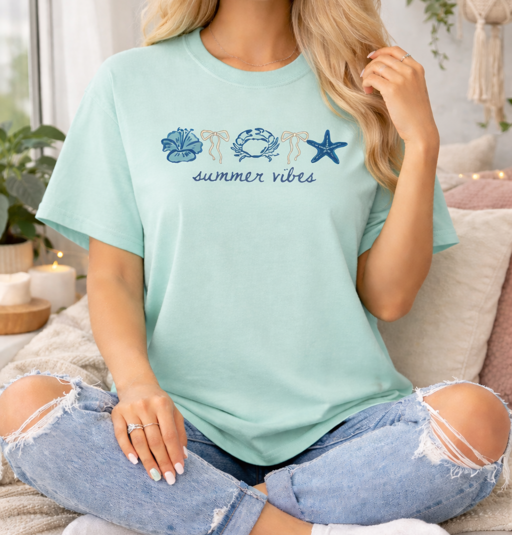 Woman wearing a light blue t-shirt with beach-themed graphics and text, sitting on a couch.